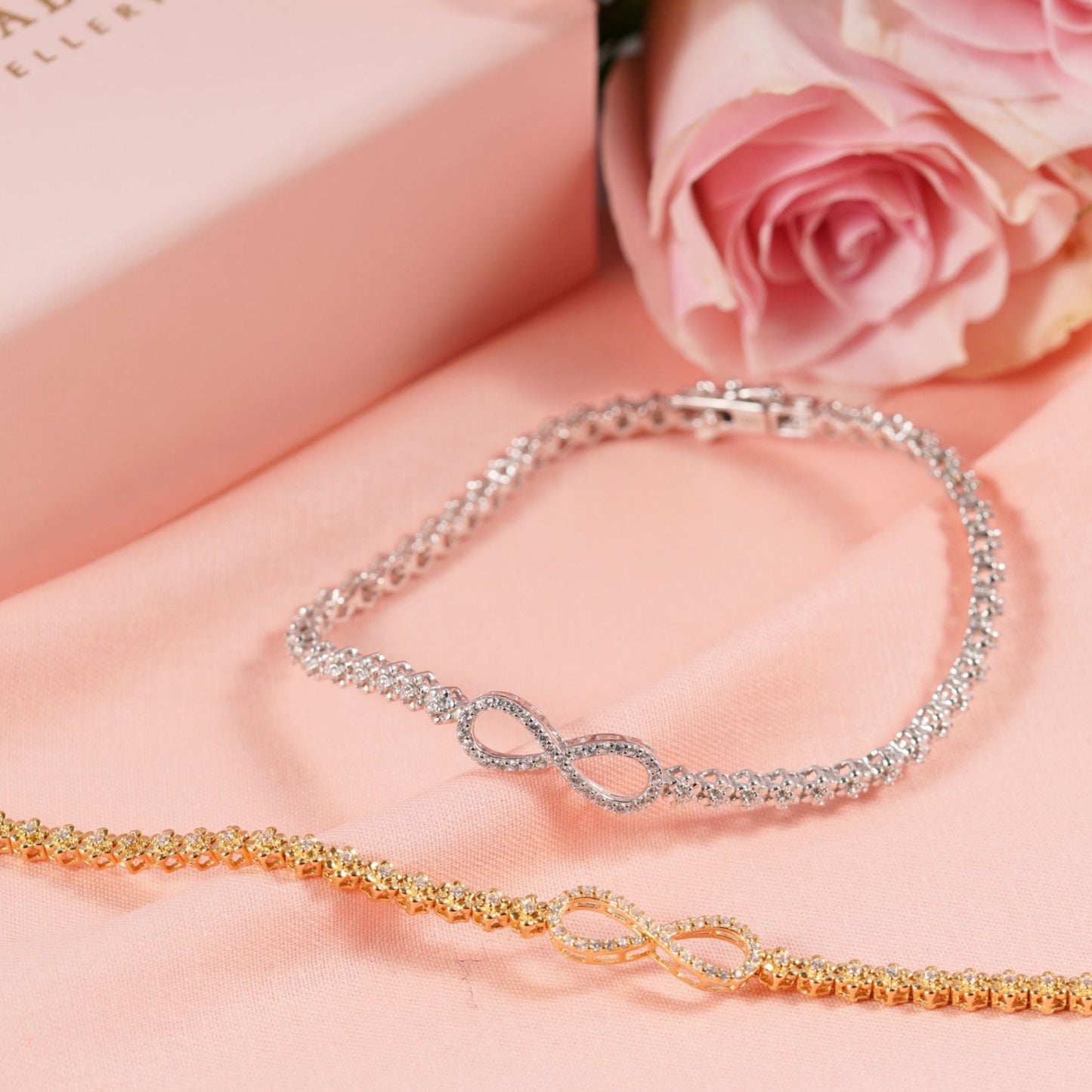 Two infinity bracelets, one gold and one silver, on a pink surface with pink roses in the background.