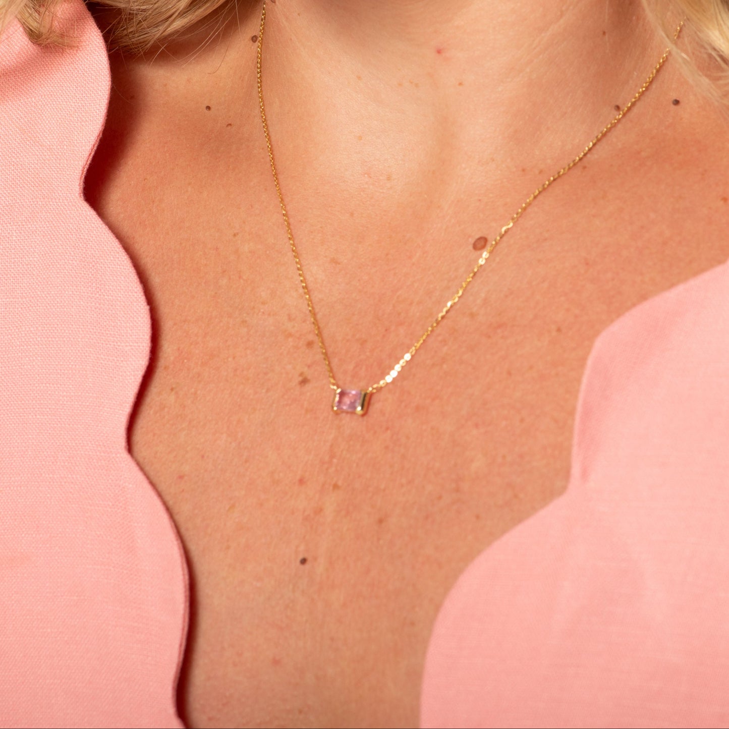 Person wearing a delicate gold necklace with a small pink pendant against a pink background