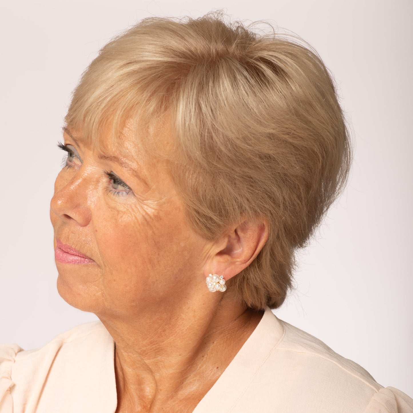 Woman with short blonde hair wearing a beige top against a white background with pearl cluster earring