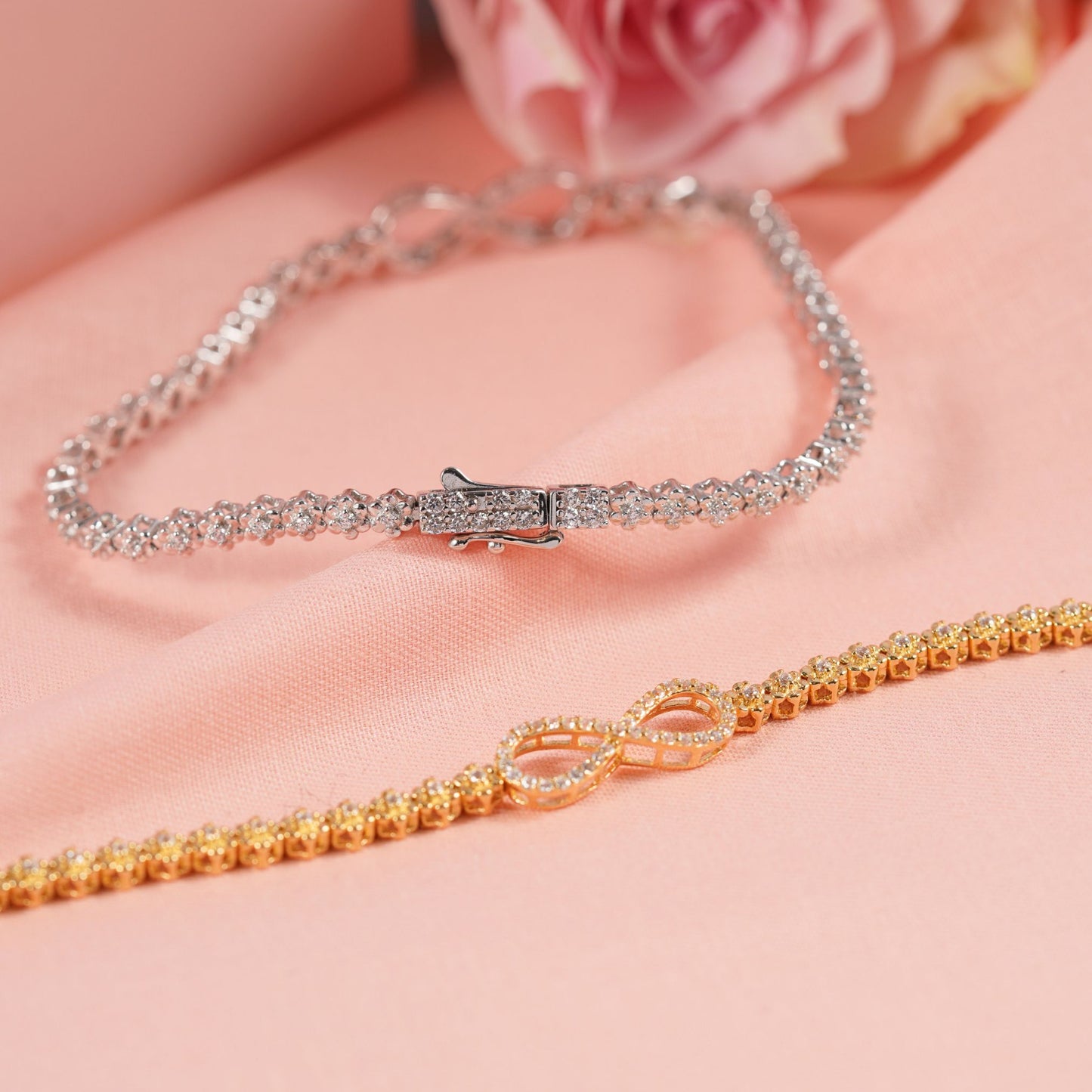 Two infinity bracelets, one gold and one silver, on a soft pink surface with a blurred floral background.