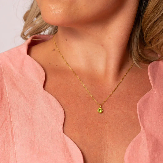 Peridot gift guide featuring a delicate gold necklace with a round green peridot pendant worn with a pink scalloped-neck blouse.