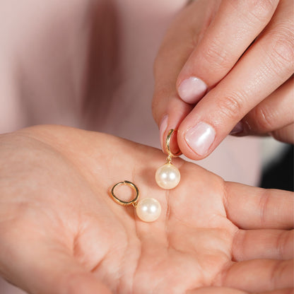 Gold huggie hoop earrings with white pearl drops held in hand